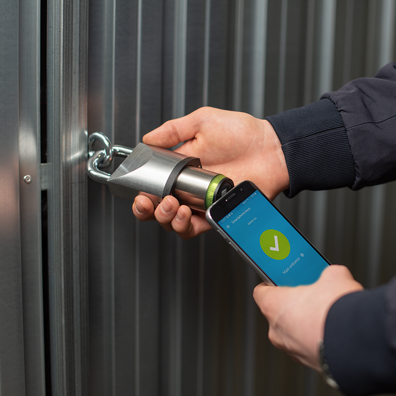 Someone holding a smartphone in front of a Tapkey Padlock. | Jemand hält ein Smartphone vor ein Tapkey-Vorhängeschloss.